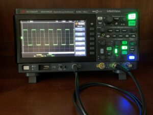 Benchtop oscilloscope displaying a green square wave on an electronics test bench